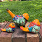 Three hand-painted ceramic bird figurines in yellow, orange  blue, red, and black, displayed on a brick surface with a green grass background left view
