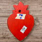 A Milagros heart featuring a gold textured top and a black base covered in raised silver-toned charms displayed on a wooden wall, back view.