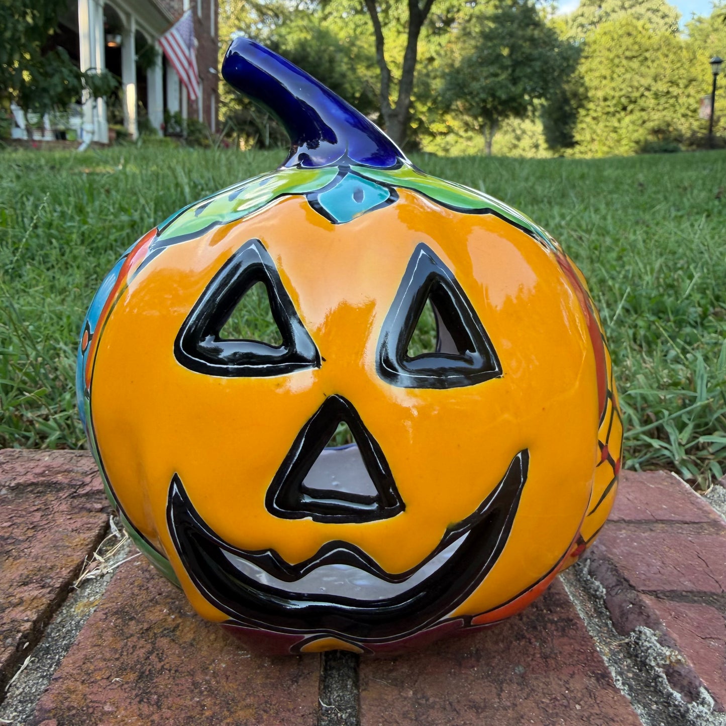 A Talavera jack-o'-lantern pumpkin decoration with a carved face, sitting on a brick surface in an outdoor yard with grass and trees in the background.