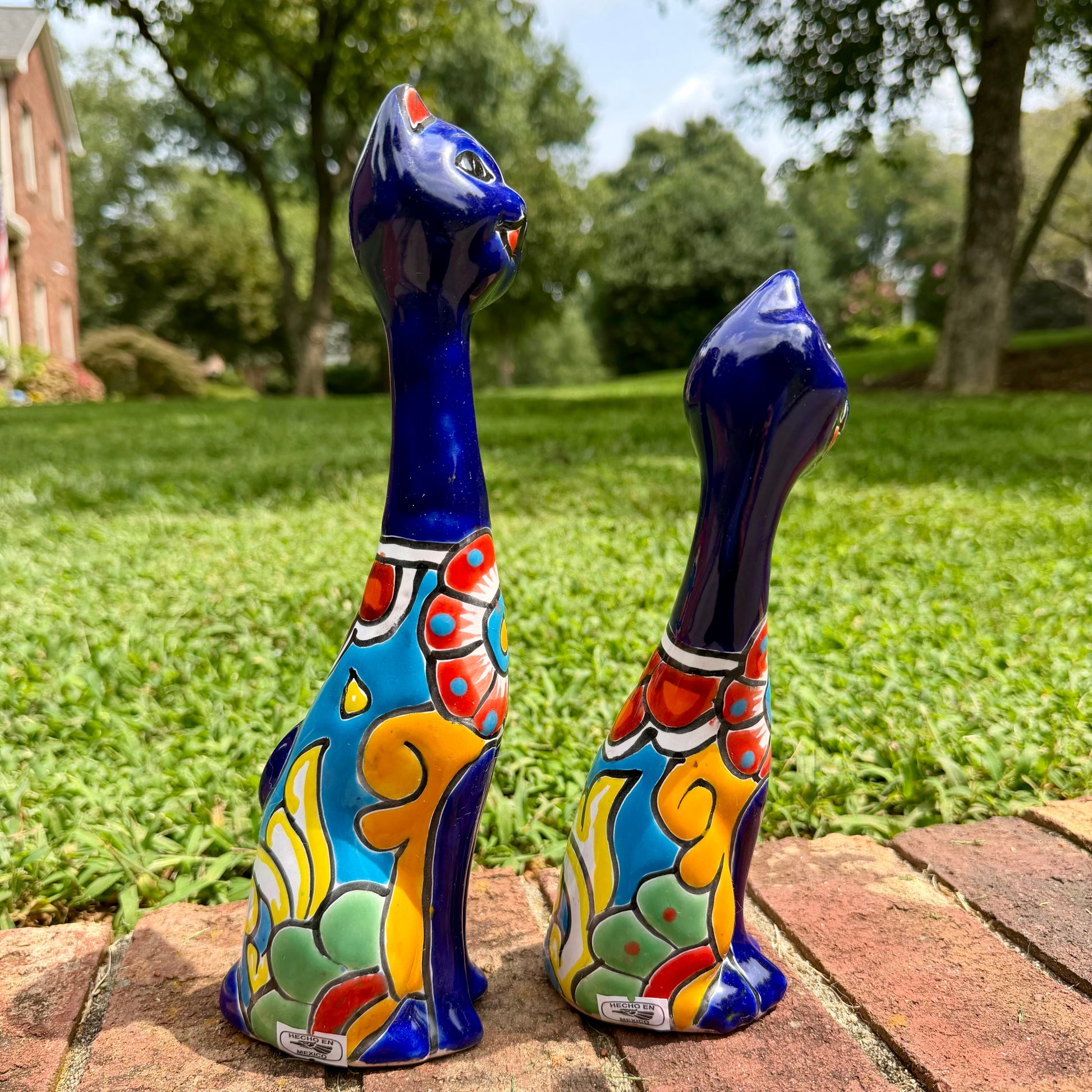 Two colorful glazed ceramic cat figurines sitting on a brick path in a grassy backyard. The cats are decorated with vibrant hand-painted patterns in blue, yellow, green, red, and white. This is their side view.