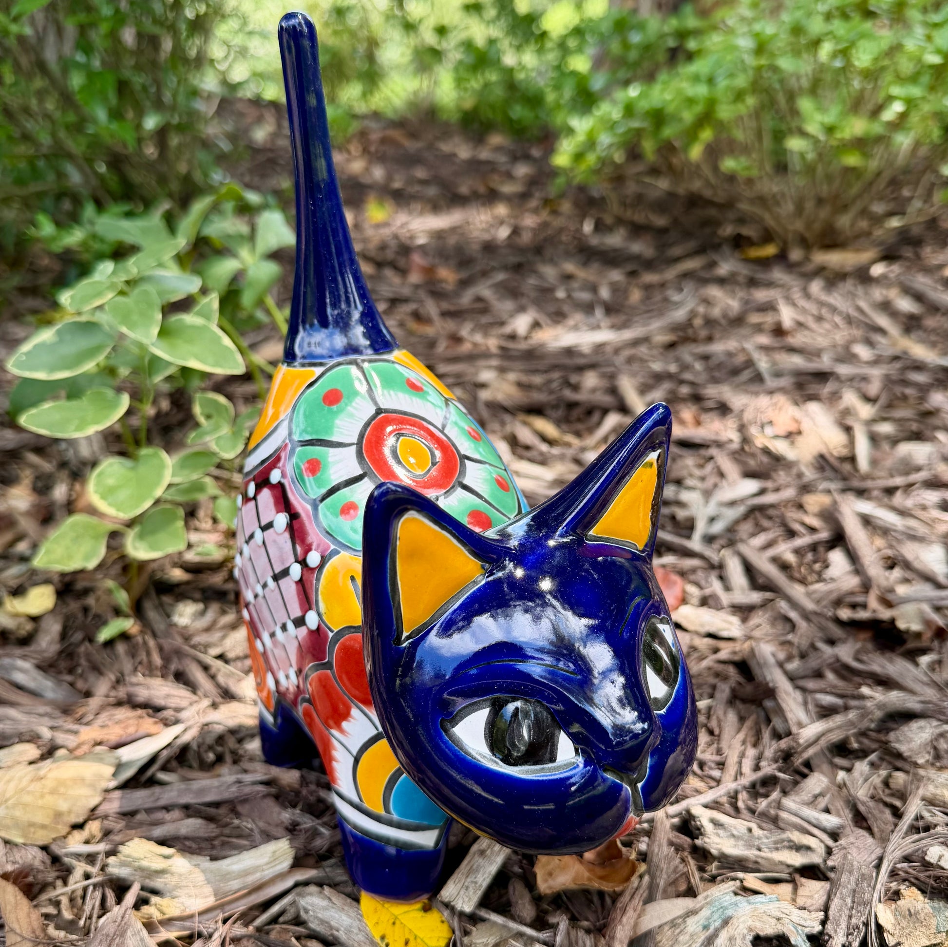 A colorful ceramic cat figurine with a glossy blue head and tail, yellow ears and paws, and a patterned body, standing outdoors on mulch with green plants in the background.