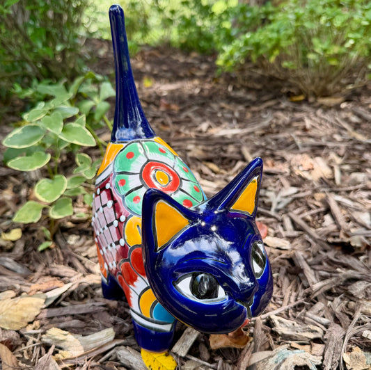 A colorful ceramic cat figurine with a glossy blue head and tail, yellow ears and paws, and a patterned body, standing outdoors on mulch with green plants in the background.
