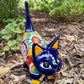 A colorful ceramic cat figurine with a glossy blue head and tail, yellow ears and paws, and a patterned body, standing outdoors on mulch with green plants in the background.