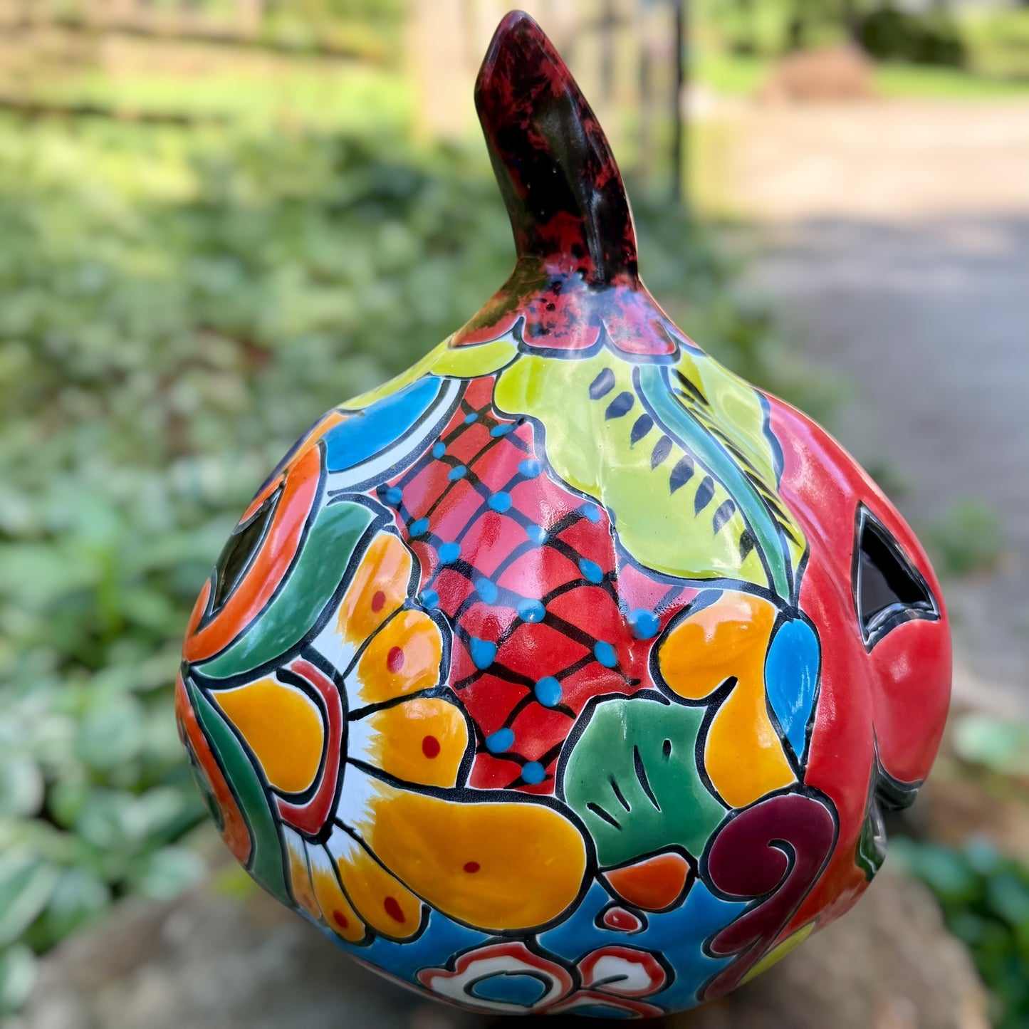 A hand-painted ceramic jack-o'-lantern pumpkin with a red face, black carved eyes and mouth, and a multicolored floral pattern on a garden background. Left side view of pumpkin.