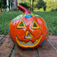 A hand-painted Talavera  jack-o'-lantern pumpkin sits on a brick patio with a green grass lawn and trees in the background. The pumpkin features a glossy orange base with hand-painted details in red, yellow, blue, and green.