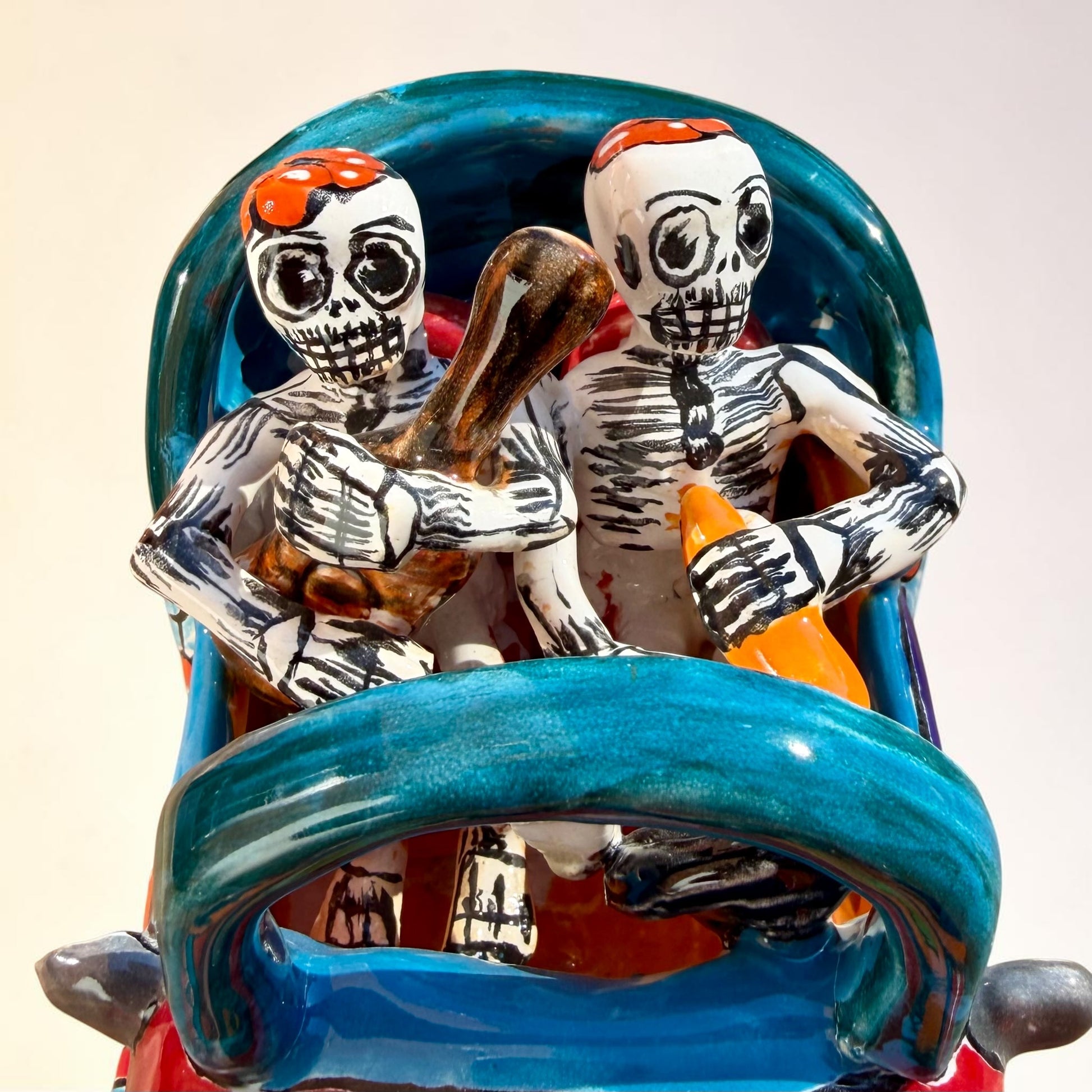 A colorful hand-painted ceramic Mexican-style convertible car figurine with Day of the Dead skeleton characters inside, shown on a neutral beige background. Close of the two catrina men.