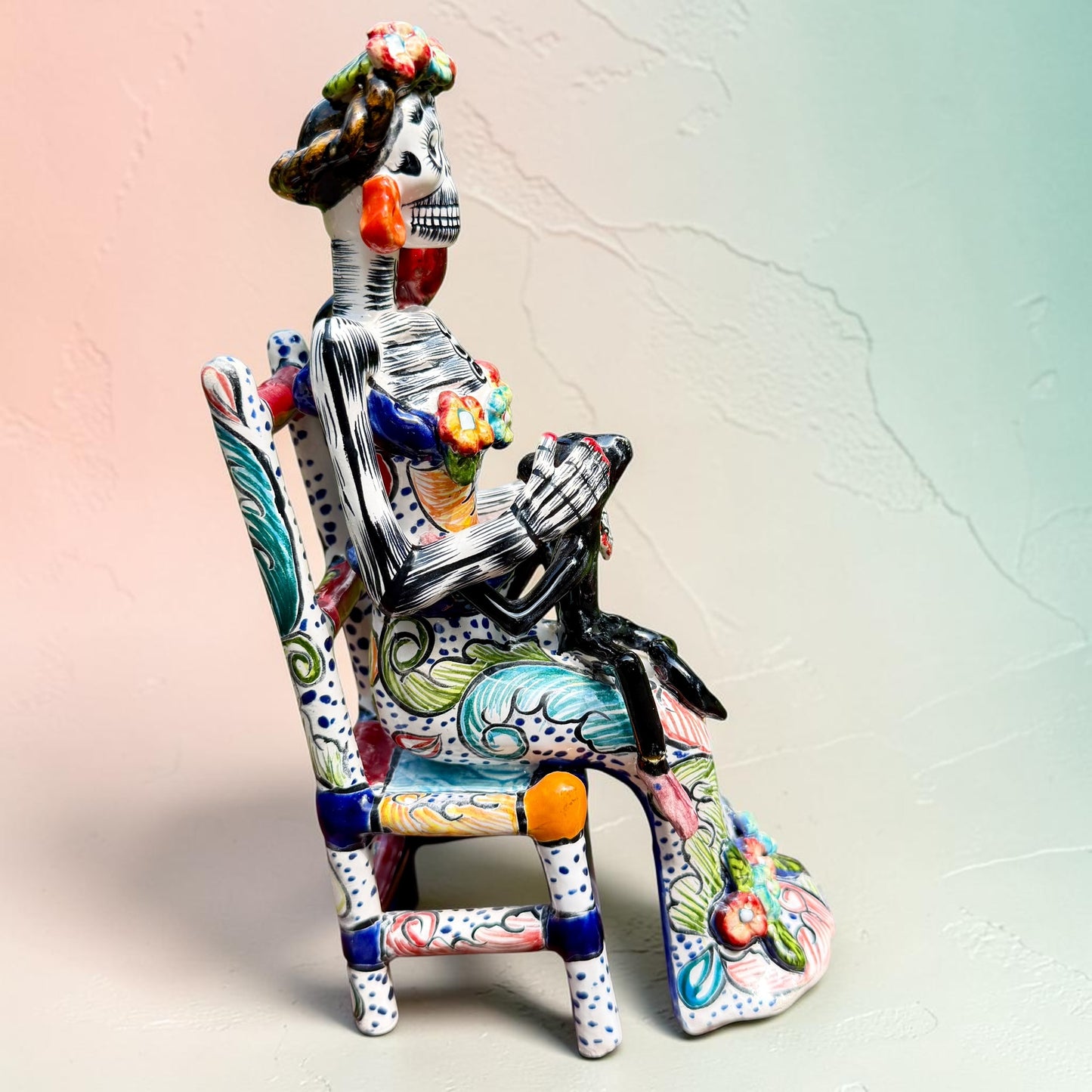 Frida Kahlo Talavera day of the dead figurine sitting in a chair holding a monkey with a parrot on her shoulder, right side view.