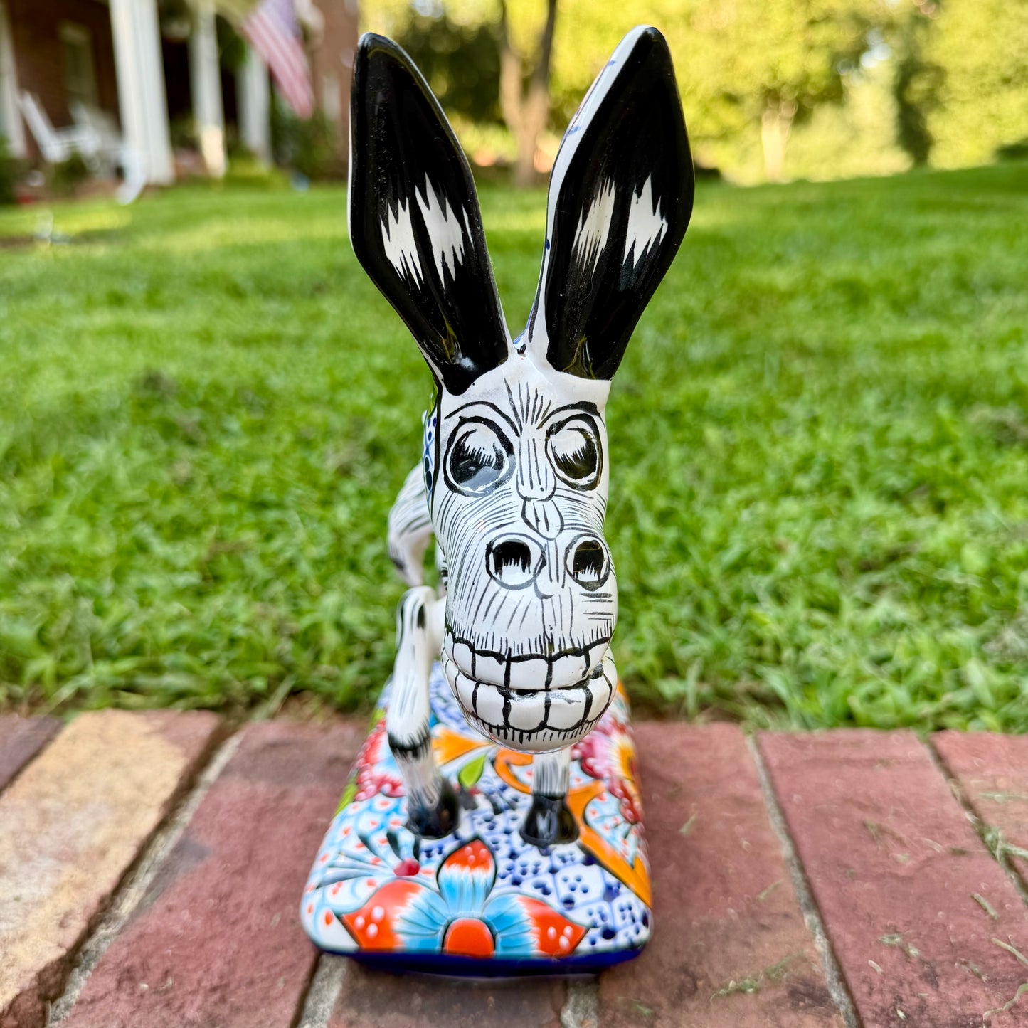 A hand-painted Mexican Talavera-style donkey figurine with a skeletal design, standing on a colorful floral base outdoors on a brick path with a green lawn in the background. This is a photo of the front facing view.