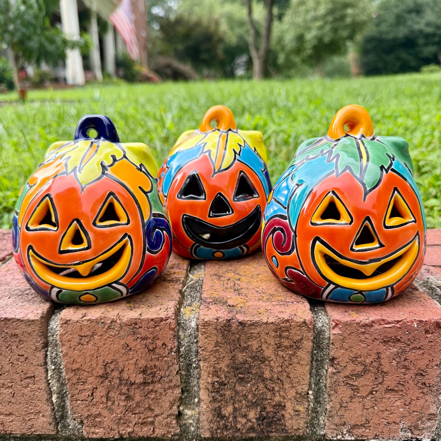 Three hand-painted talavera ceramic orange face jack-o'-lantern lanterns in vibrant orange, yellow, blue, green, and red, displayed on a brick ledge with a blurred green lawn and trees in the background.