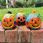 Three hand-painted talavera ceramic orange face jack-o'-lantern lanterns in vibrant orange, yellow, blue, green, and red, displayed on a brick ledge with a blurred green lawn and trees in the background.