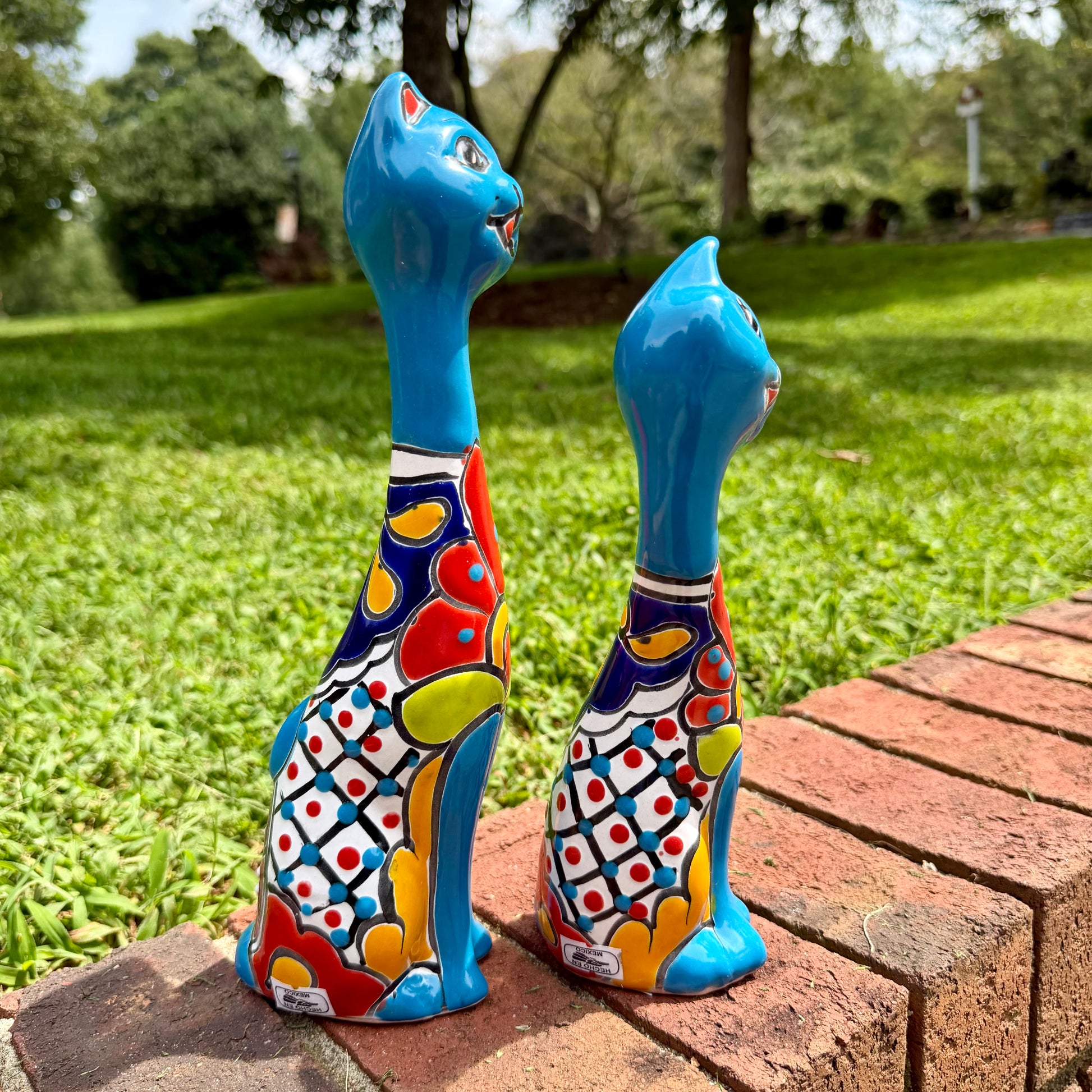 Two colorful hand-painted ceramic cat figurines sitting on a brick ledge in a grassy backyard. This is their left side view.