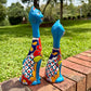 Two colorful hand-painted ceramic cat figurines sitting on a brick ledge in a grassy backyard. This is their left side view.