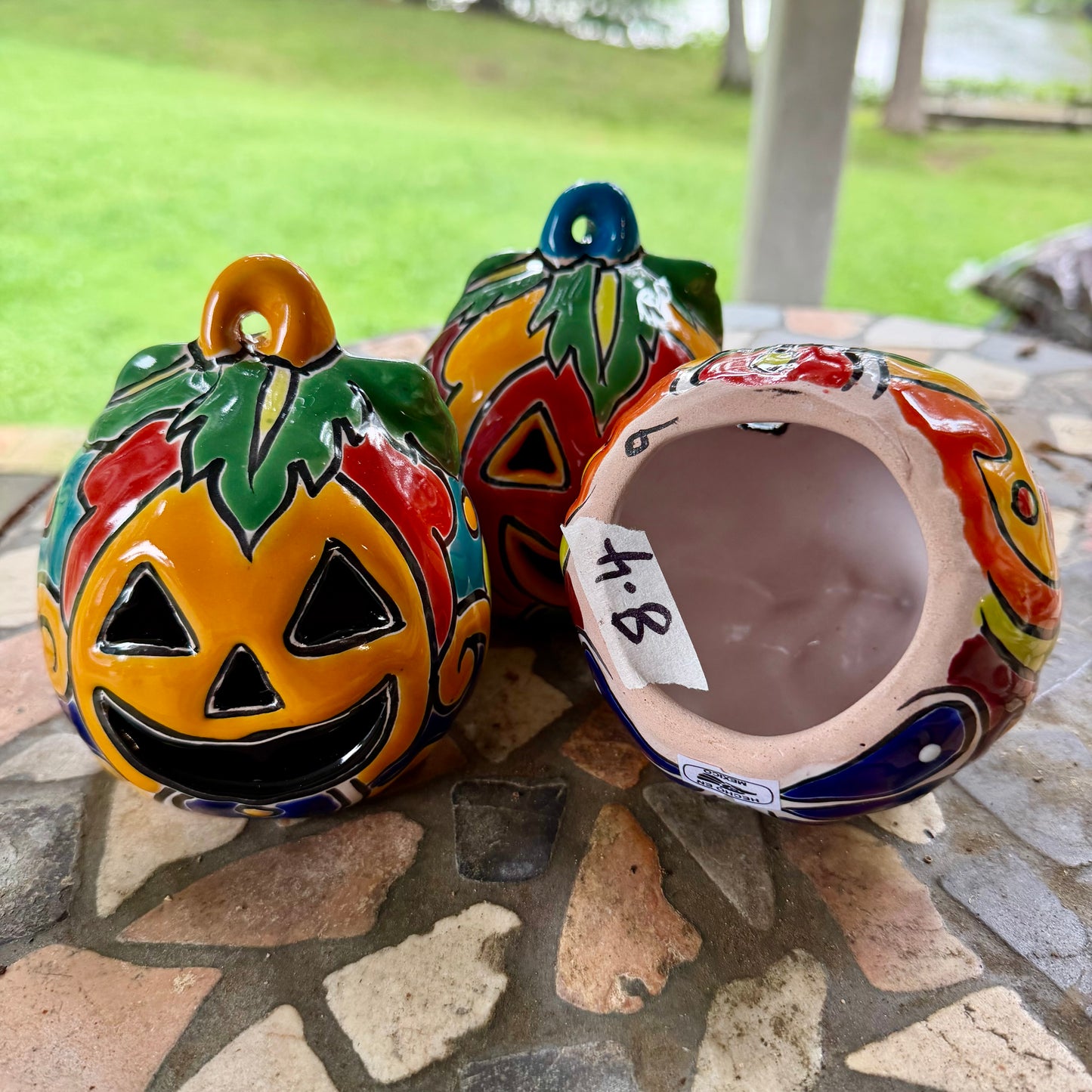 Three colorful hand-painted Talavera jack-o'-lantern pumpkins with multi-color displayed on a stone patio with a blurred green lawn background, bottom view.