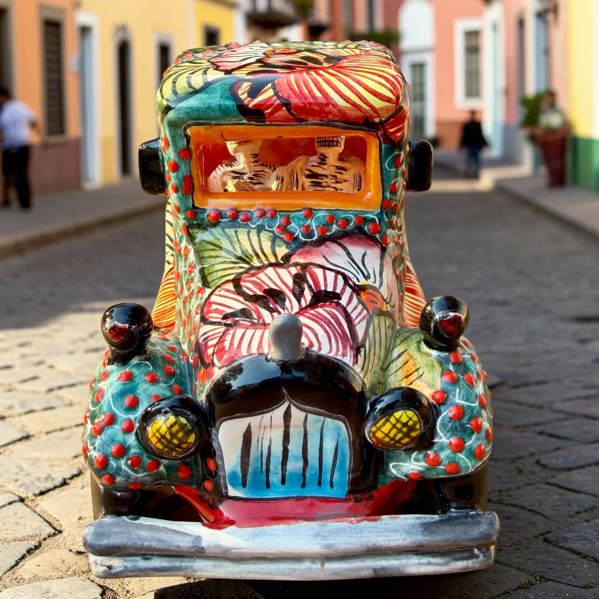 A decorative Talavera day of the dead pick up truck figurine two skeleton passengers. The truck is vibrant, with a hand-painted tropical floral pattern, photographed outdoors on a stone-paved street. This is a photo of the front of the pick up truck.