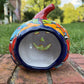 A hand-painted Talavera  jack-o'-lantern pumpkin sits on a brick patio with a green grass lawn and trees in the background. The pumpkin features a glossy orange base with hand-painted details in red, yellow, blue, and green. Back view with opening for candle.