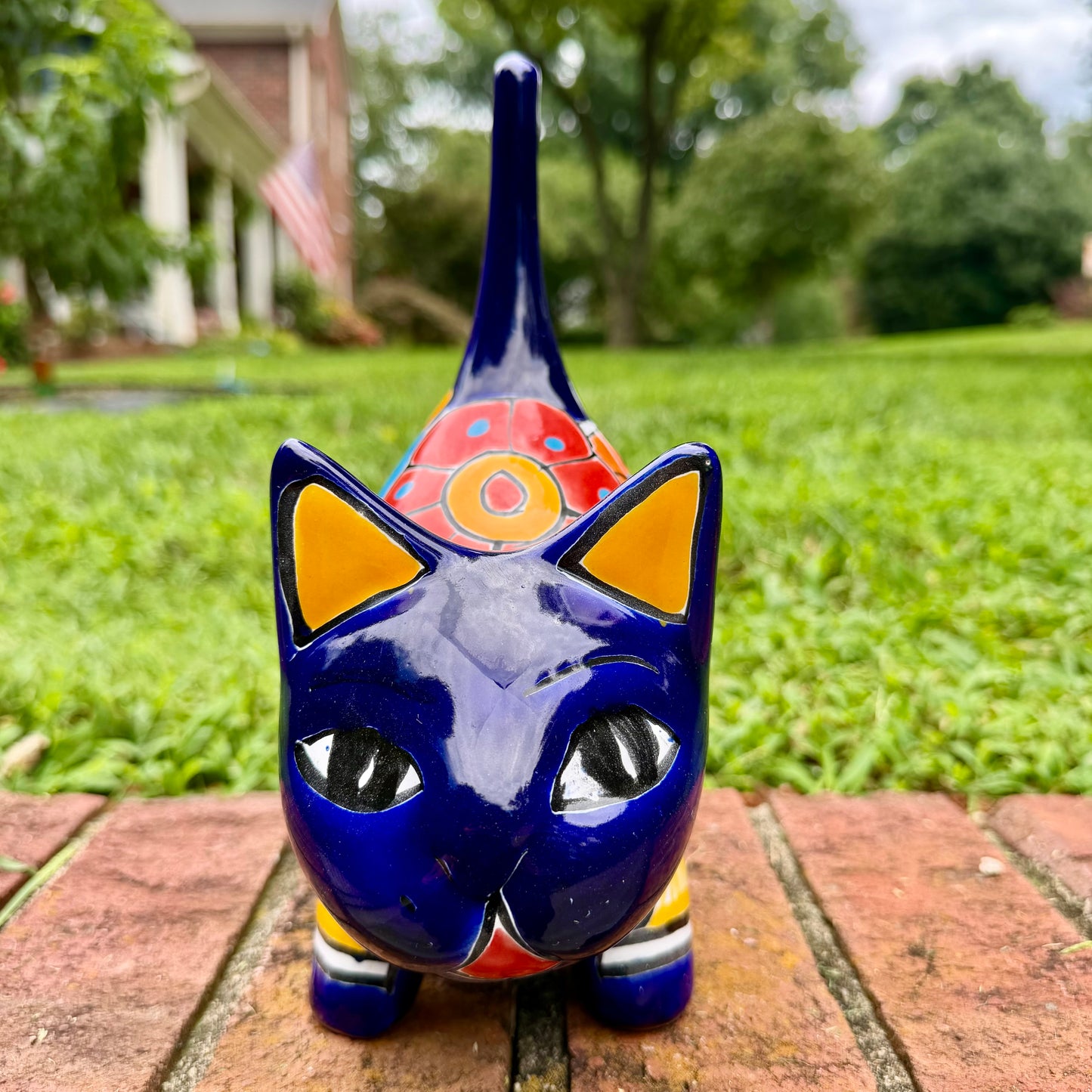 A glossy talavera ceramic cat figurine decorated with vibrant hand-painted patterns in blue, orange, yellow, and red, standing on a brick path with a green lawn and a house in the blurred background. Front face view.