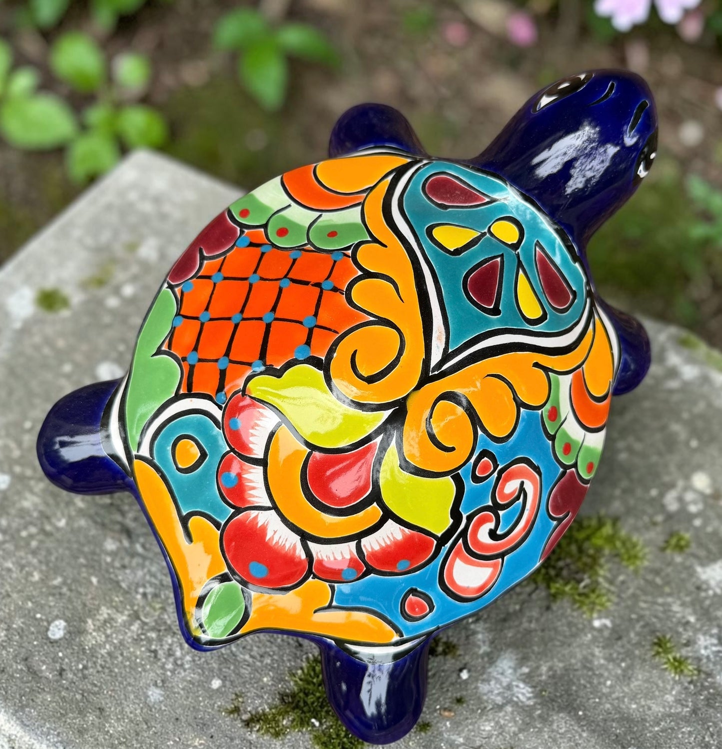 A glossy ceramic turtle figurine with a vibrant hand-painted patterned shell and a deep blue head and legs, resting on a gray stone surface with a blurred green garden background. Top view.