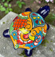 A glossy ceramic turtle figurine with a vibrant hand-painted patterned shell and a deep blue head and legs, resting on a gray stone surface with a blurred green garden background. Top view.