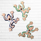 Three colorful ceramic wall-mounted frog sculptures in a vibrant multicolor design, displayed against a white brick wall background back view showing hanging hole.