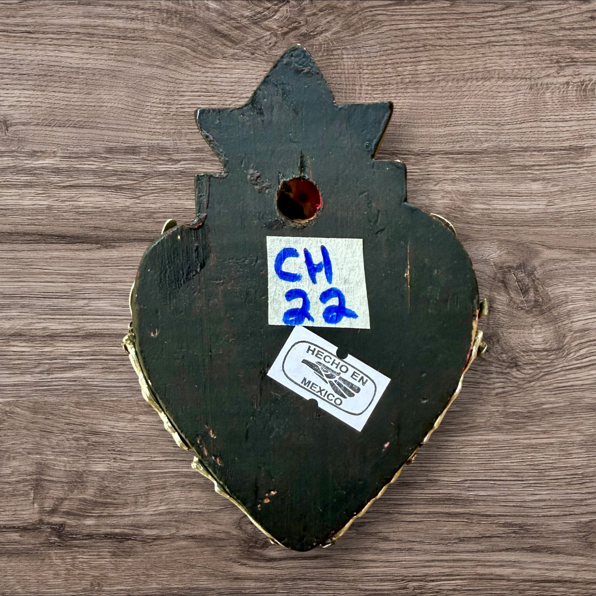 A decorative Milagros Sacred Heart featuring a black and silver painted heart adorned with charms, set against a rustic wood background, this is the back view.