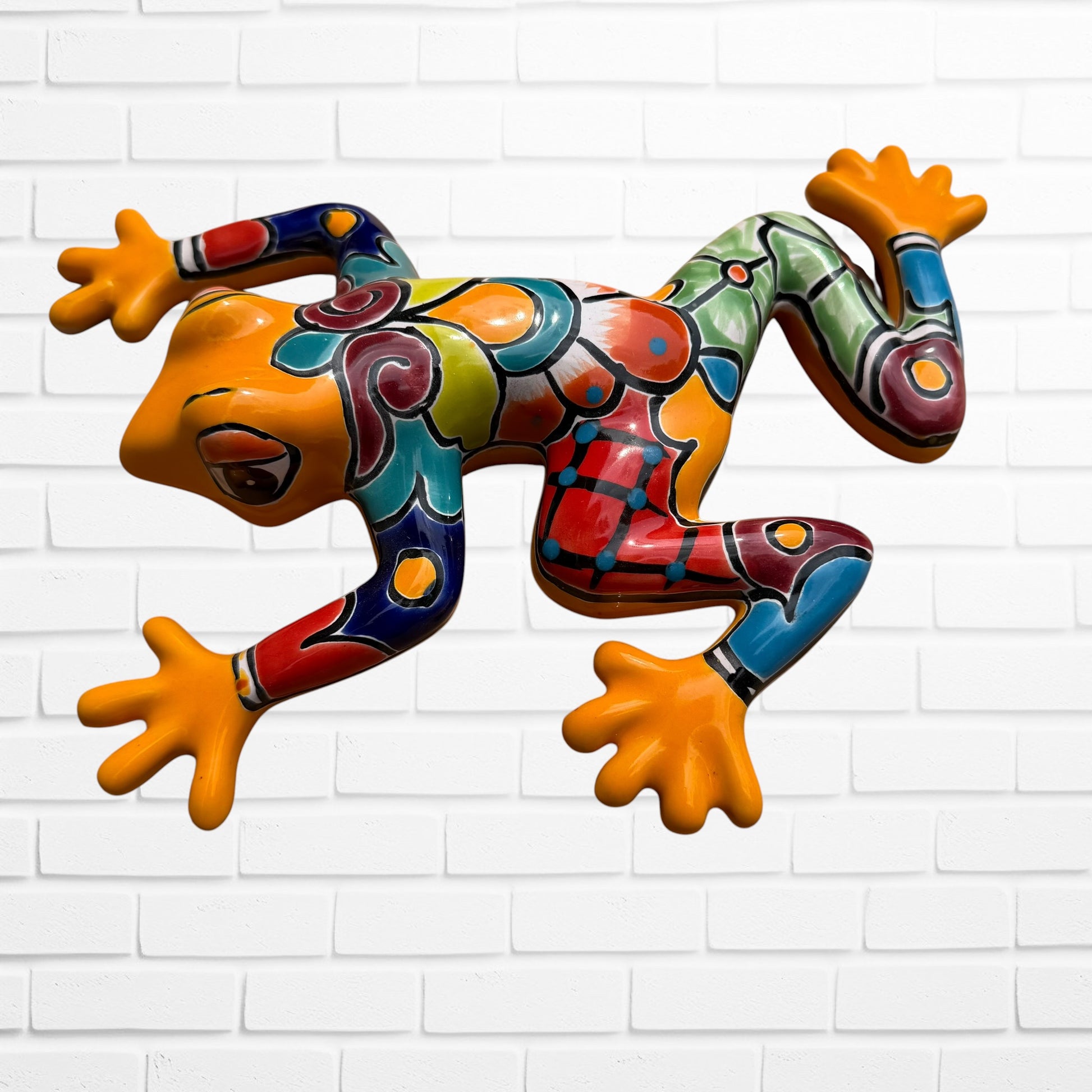 A colorful hand-painted ceramic frog wall sculpture in a dynamic leaping pose, set against a white painted brick wall background.