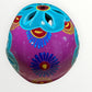 A hand-painted ceramic sugar skull in vibrant purple, turquoise blue, and yellow, decorated with floral and geometric patterns on a plain white background skull back view.