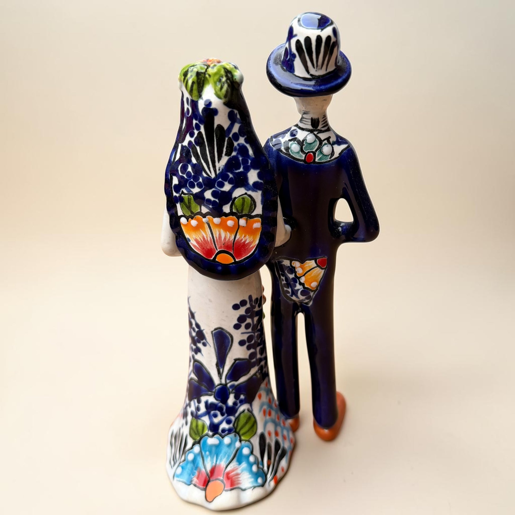 A hand-painted Talavera Day of the Dead bride and groom figurine. The groom is dressed in a blue suit with white patterns, a black bow tie, and an orange-red bow tie accent, wearing a blue hat. Back view of couple showing veil and tuxedo tail.