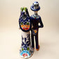 A hand-painted Talavera Day of the Dead bride and groom figurine. The groom is dressed in a blue suit with white patterns, a black bow tie, and an orange-red bow tie accent, wearing a blue hat. Back view of couple showing veil and tuxedo tail.
