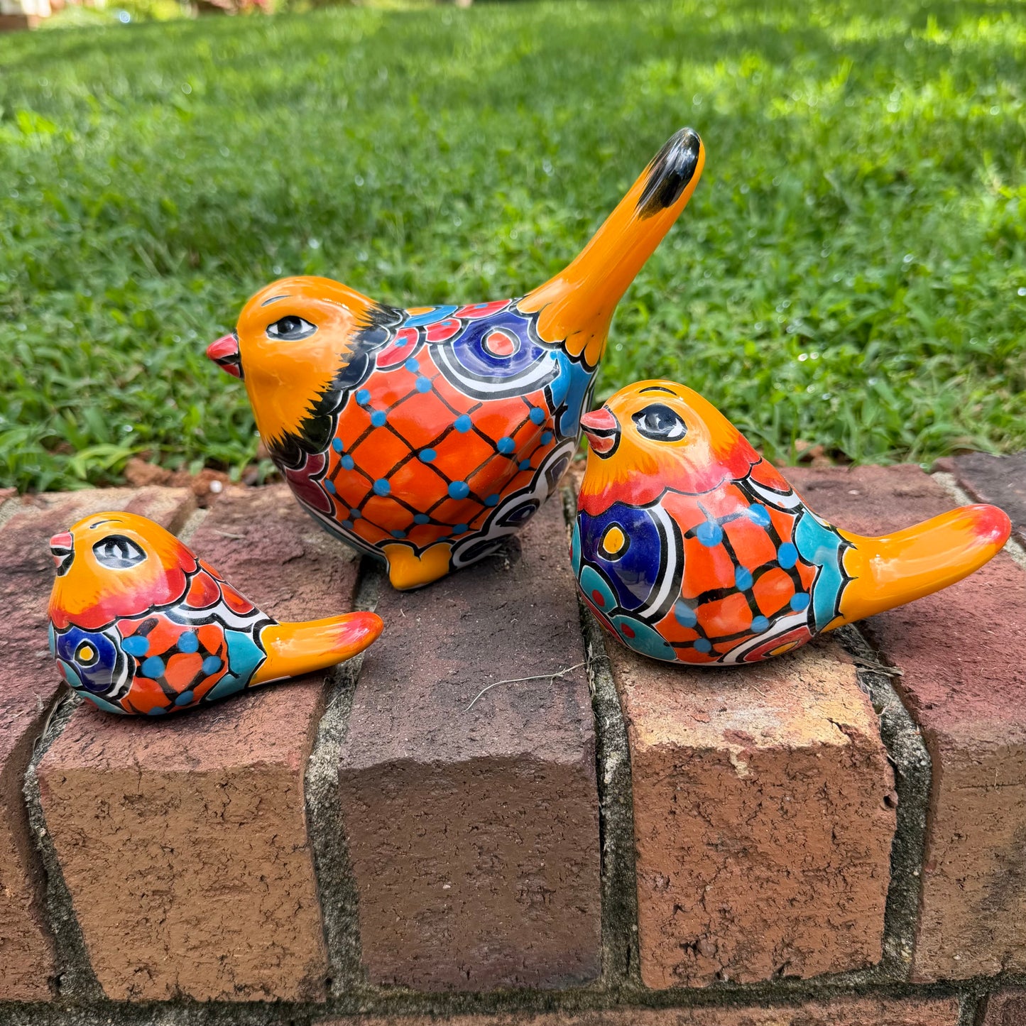 Three hand-painted ceramic bird figurines in yellow, orange  blue, red, and black, displayed on a brick surface with a green grass background. right side view.