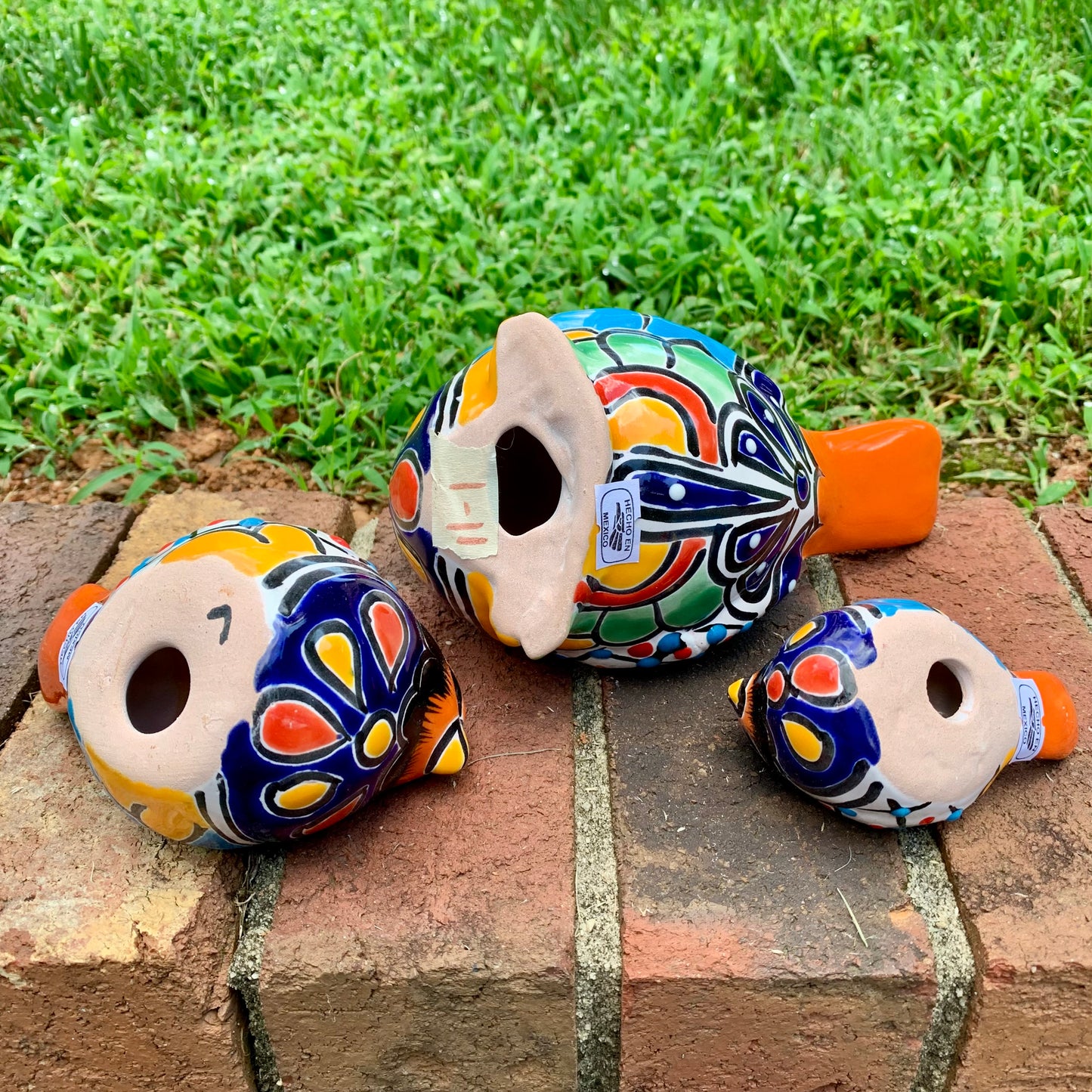 Three hand-painted ceramic bird garden statues in orange, blue, yellow, and black, displayed on a brick ledge in a backyard with a green lawn and trees in the background bottom figurine view.