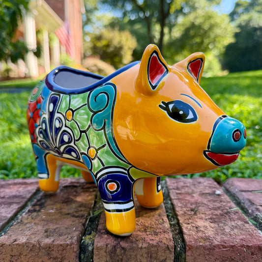 A colorful ceramic pig planter with a yellow-orange face and body, decorated with blue, green, red, and white floral patterns, sitting on a brick surface with a blurred outdoor garden background.