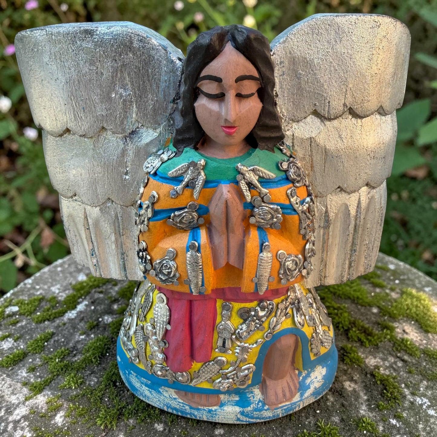 Milagros wooden angel figurine with silver wings and metallic charms, displayed outdoors on a mossy stone with green foliage in the background. The angel is in preyer.