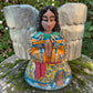 Milagros wooden angel figurine with silver wings and metallic charms, displayed outdoors on a mossy stone with green foliage in the background. The angel is in preyer.