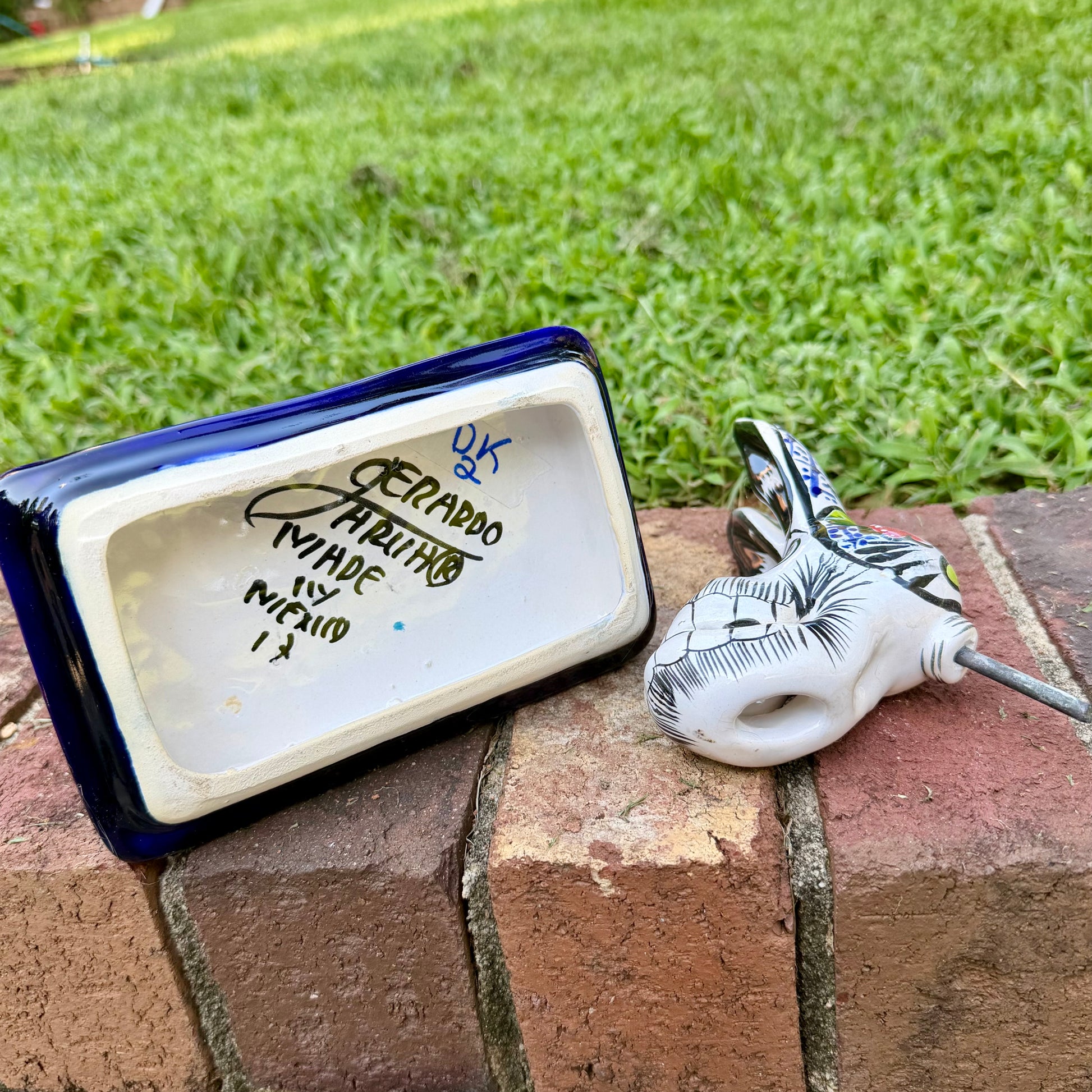 A colorful ceramic donkey Catrina figurine with a skeletal white-and-black design and a floral base, photographed outdoors on a brick surface with a blurred green lawn and house in the background. Bottom view with artist signature.
