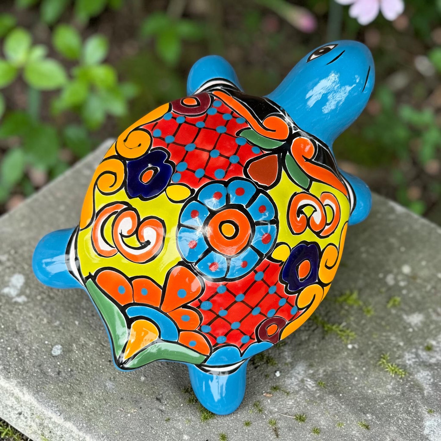A hand-painted ceramic turtle figurine resting on a mossy stone outdoors, featuring a bright blue body and a highly decorated shell with vibrant orange, yellow, red, and blue patterns, top view of Shell.
