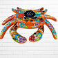 A colorful talavera crab wall art sculpture decorated with vibrant floral patterns, shown against a white brick wall background, smiling face view.