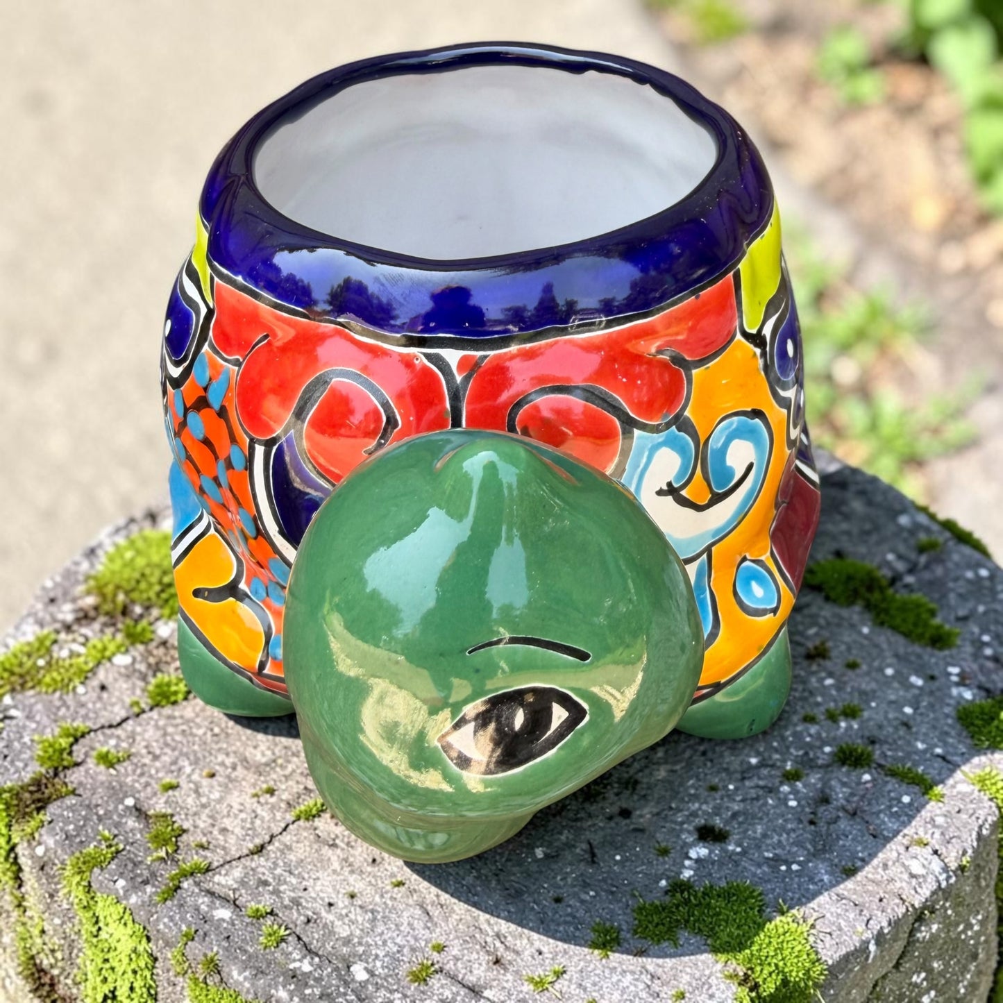 A colorful ceramic turtle-shaped planter with a green head and legs, decorated with vibrant hand-painted patterns in orange, yellow, blue, red, and white, sitting on a mossy stone outdoors. Front of turtle by neck and head.
