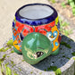 A colorful ceramic turtle-shaped planter with a green head and legs, decorated with vibrant hand-painted patterns in orange, yellow, blue, red, and white, sitting on a mossy stone outdoors. Front of turtle by neck and head.
