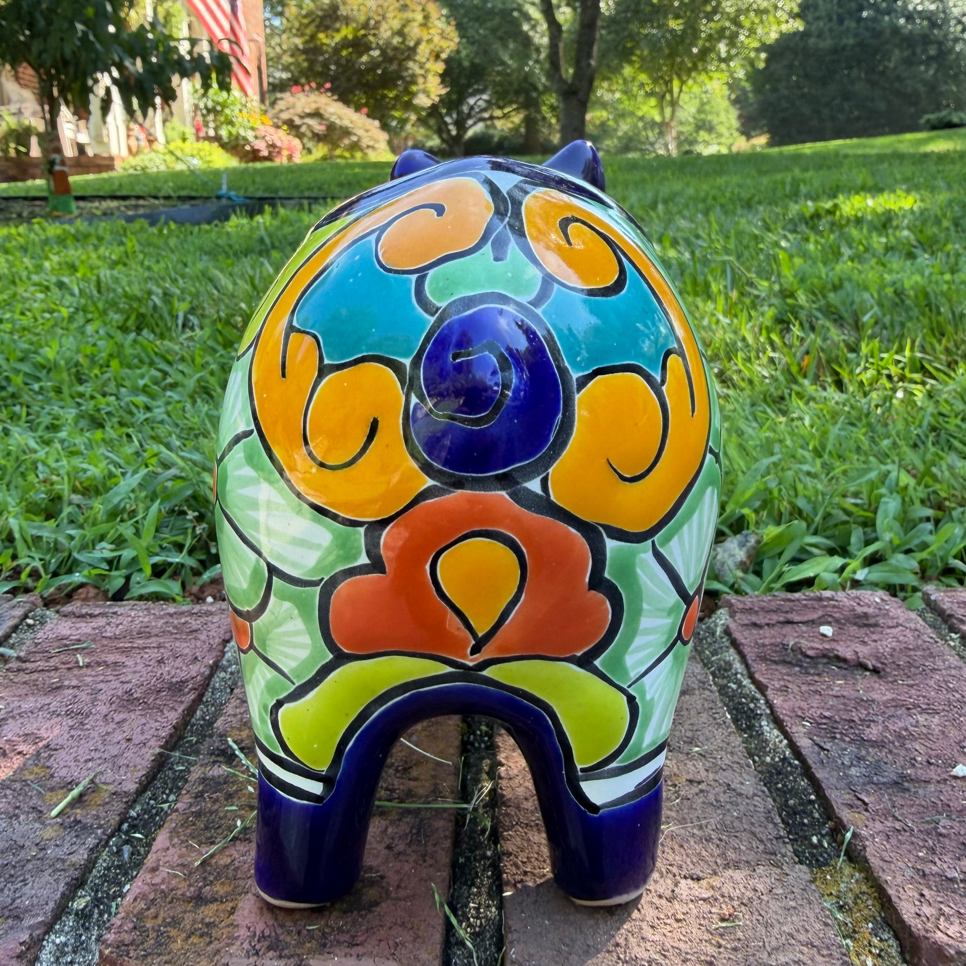 A glossy talavera pig figurine decorated with vibrant hand-painted patterns in blue, yellow, red, green, and white, standing on a brick surface with a blurred outdoor garden background, back tail view.