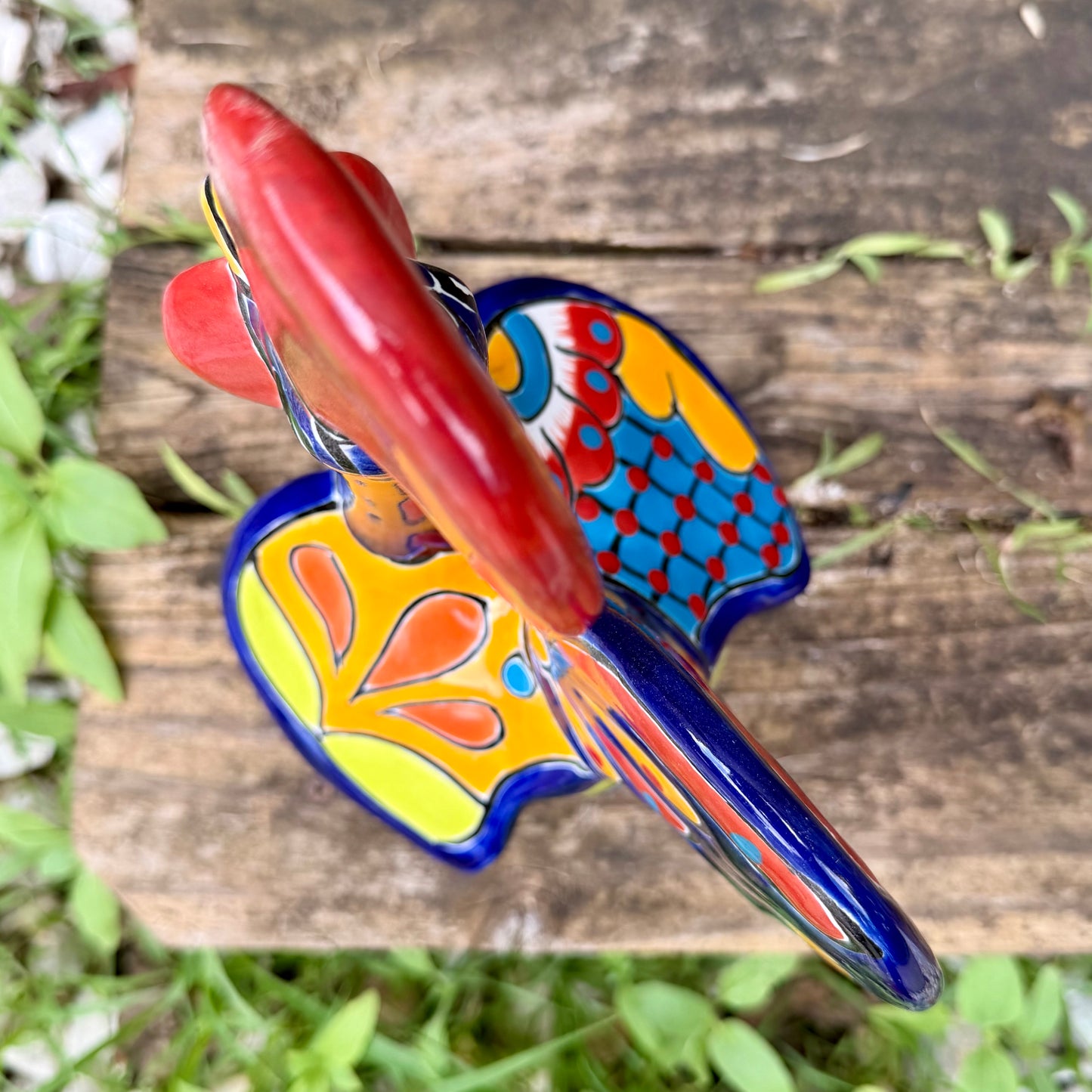 A colorful hand-painted ceramic rooster figurine standing on a rustic wooden surface in front of a green wooden door, top view.