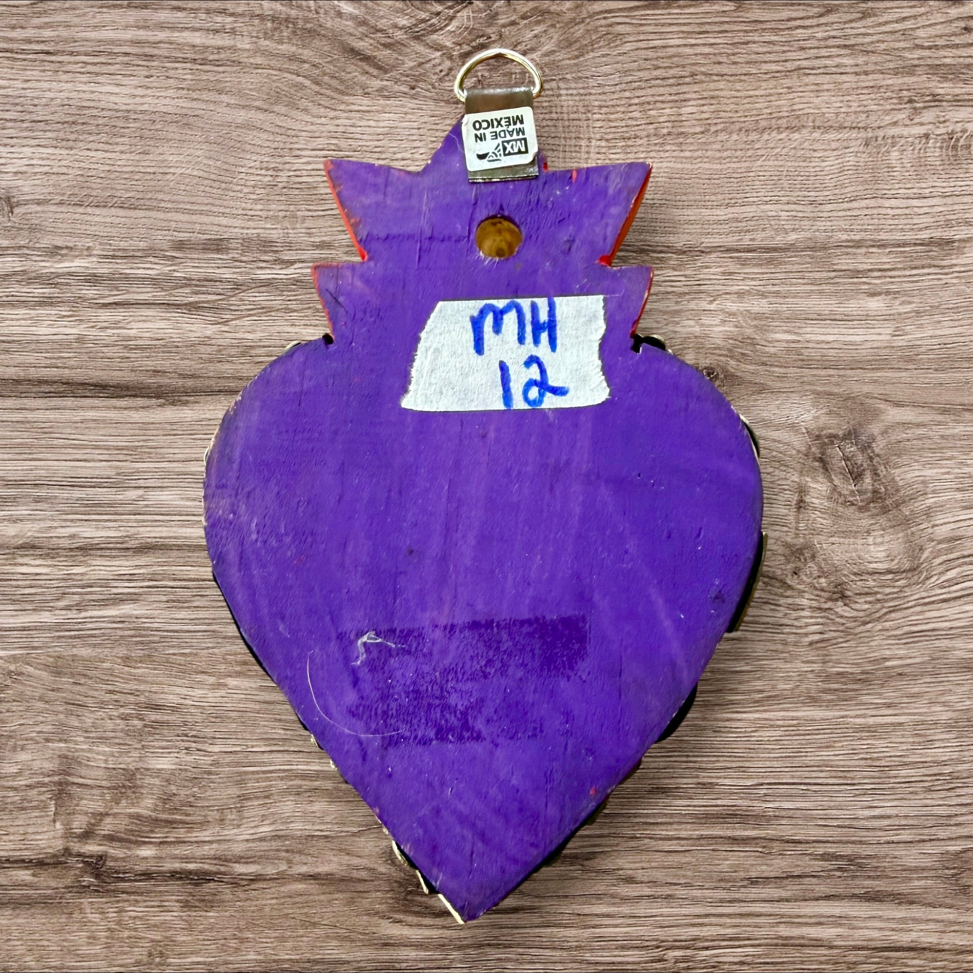 A Milagros heart featuring a gold textured top and a purple base covered in raised silver-toned charms displayed on a wooden wall.