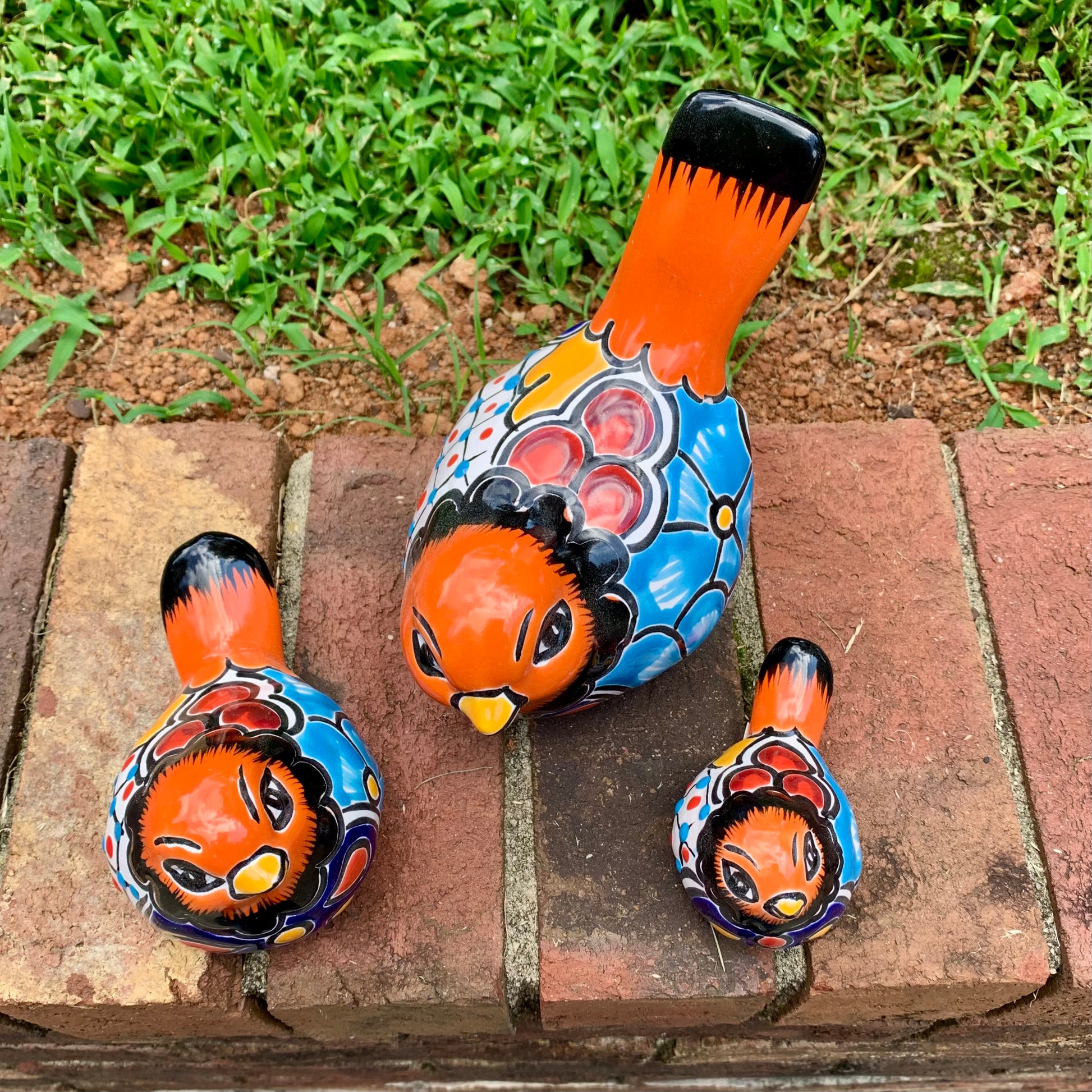 Three hand-painted ceramic bird garden statues in orange, blue, yellow, and black, displayed on a brick ledge in a backyard with a green lawn and trees in the background top view.
