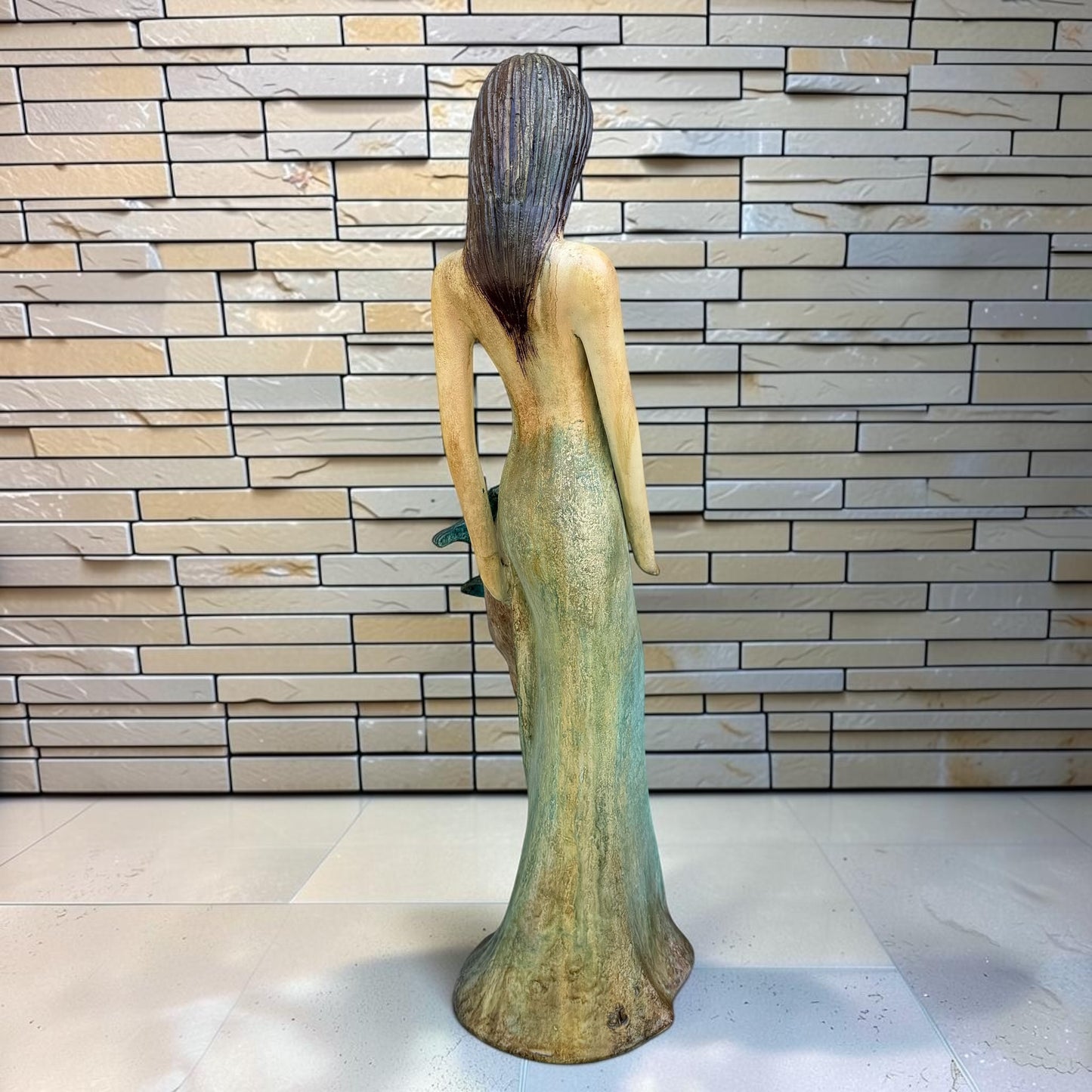 A Loretta Bertoli statue of a faceless woman in a long flowing dress with two hummingbirds, displayed against a stacked stone tile wall on a light tiled floor. Left back angle view of dress 