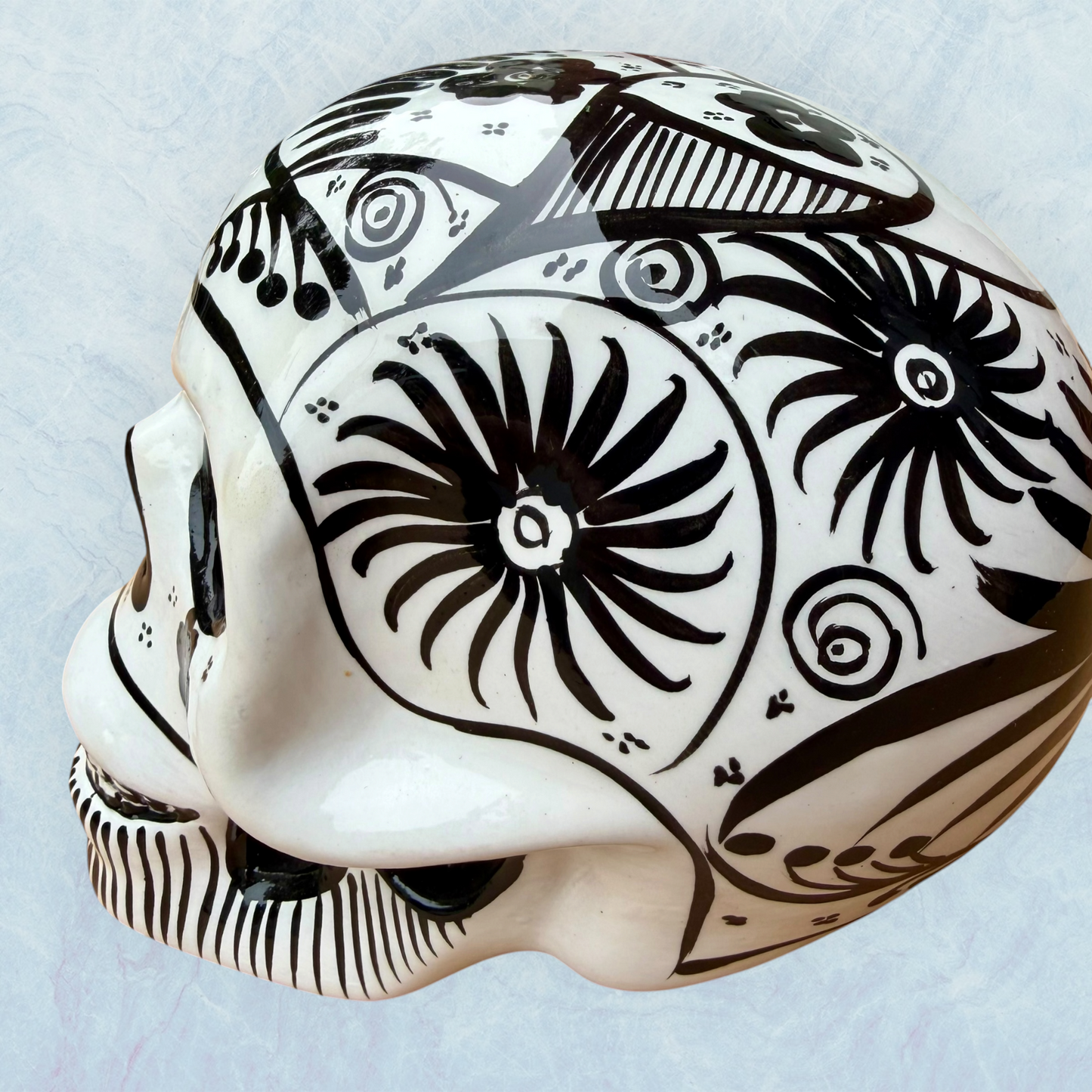 A decorative skull sculpture with intricate black sugar skull style patterns painted on an off-white base, set against a light blue textured background, right side view.