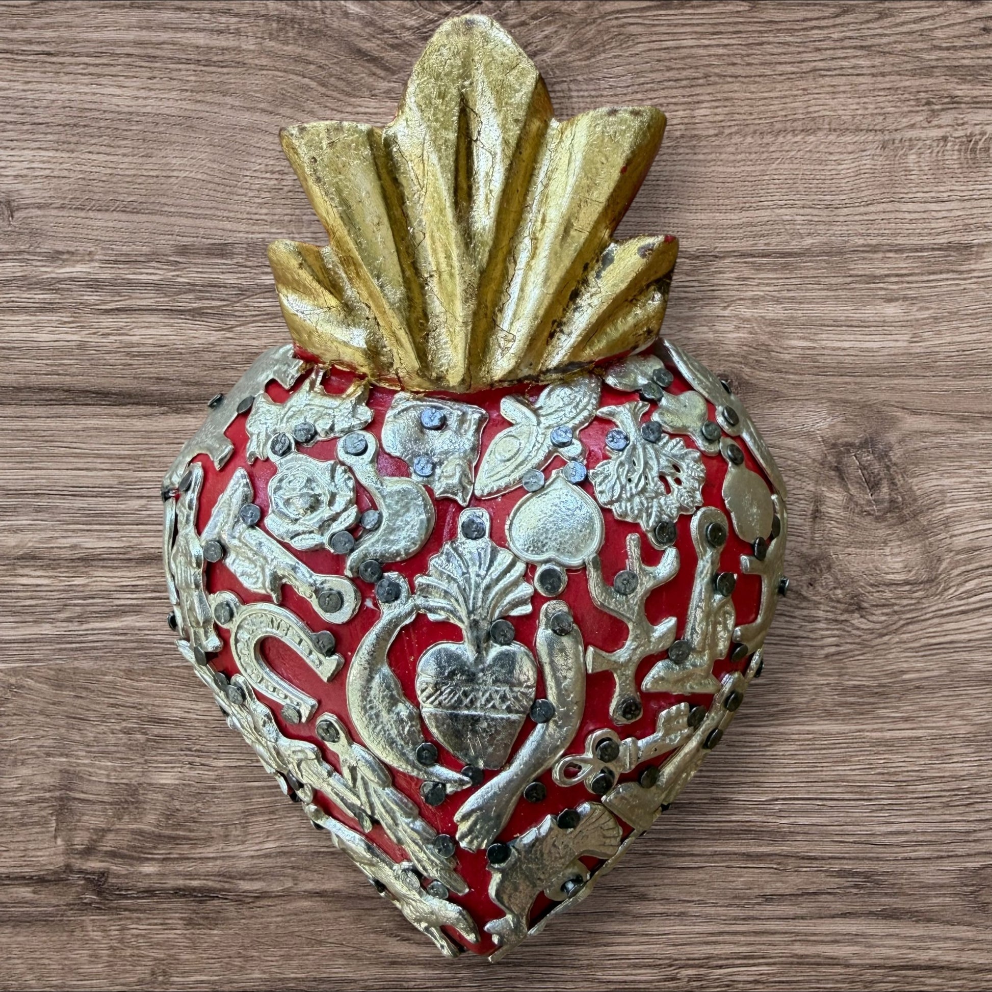 A Milagros heart featuring a gold textured top and a red heart base covered in raised silver-toned charms displayed on a wooden wall.