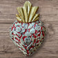 A Milagros heart featuring a gold textured top and a red heart base covered in raised silver-toned charms displayed on a wooden wall.