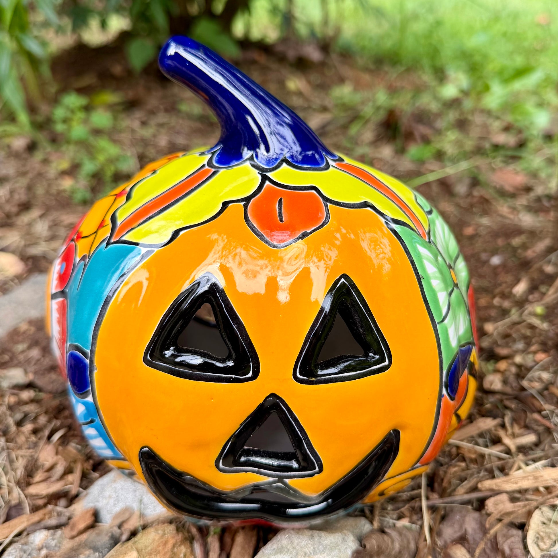 A colorful hand-painted ceramic jack-o'-lantern garden ornament with a blue stem, photographed outdoors on mulch with green foliage in the background.