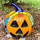 A colorful hand-painted ceramic jack-o'-lantern garden ornament with a blue stem, photographed outdoors on mulch with green foliage in the background.