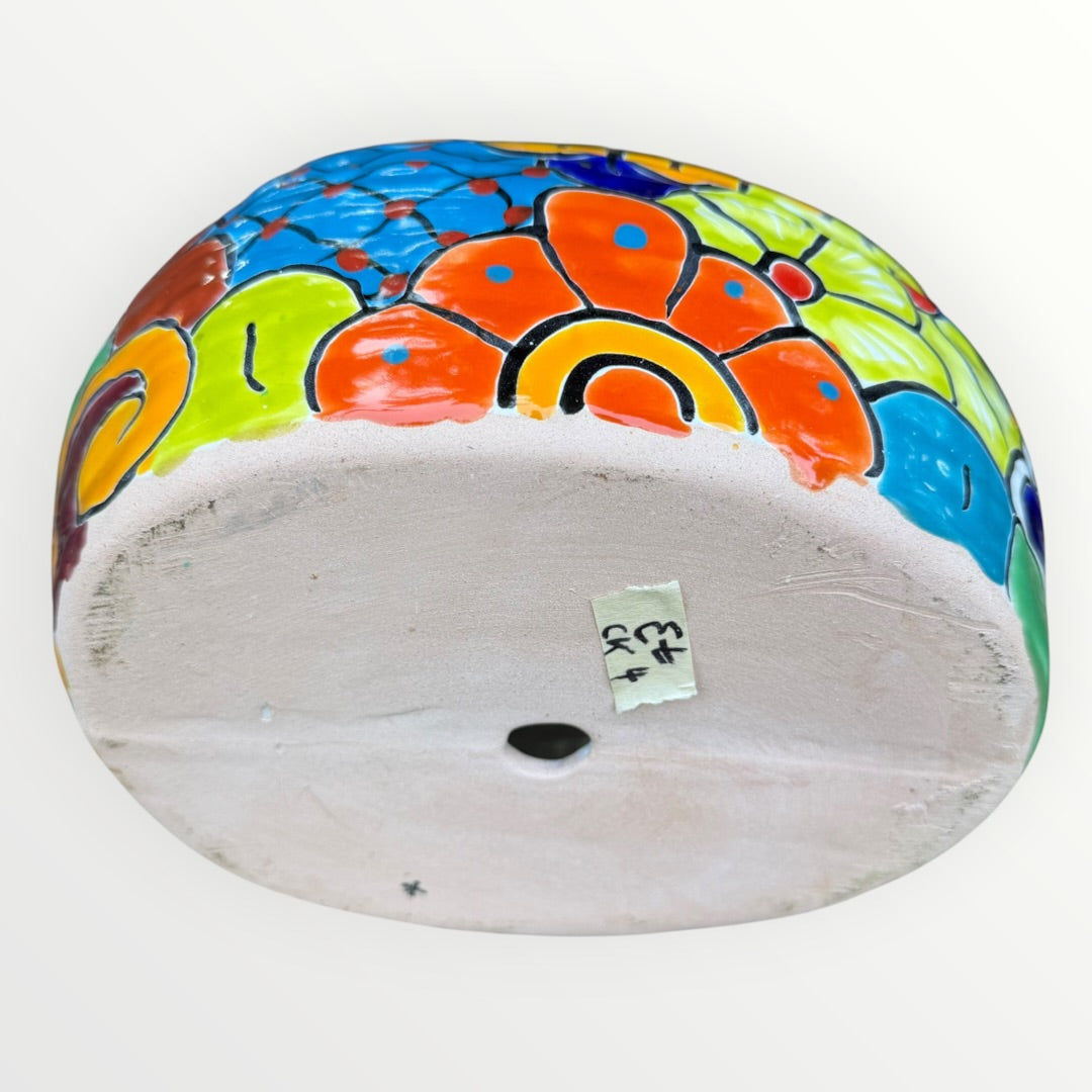 A colorful ceramic chicken-shaped planter with a glossy hand-painted finish, placed outdoors on a bed of small gray rocks and greenery, bottom view with planting hole.