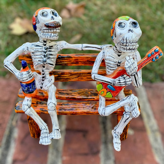 Two skeleton figurines sitting on a bench with one playing a guitar and the other holding a bottle.